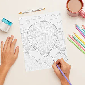 A hand colors a page of hot air balloons over the mountains.