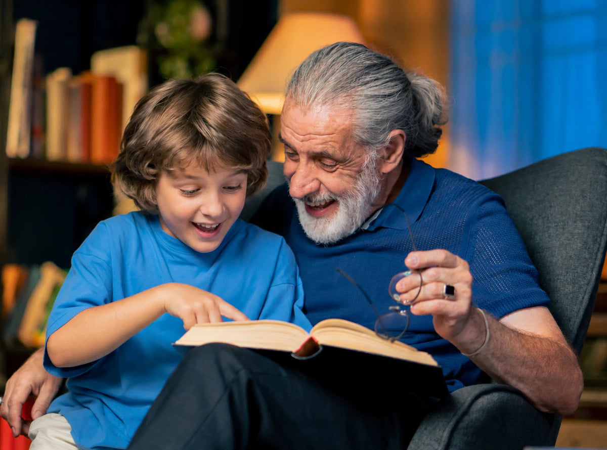 A grandfather and his grandson laugh over a book together in a cozy armchair.