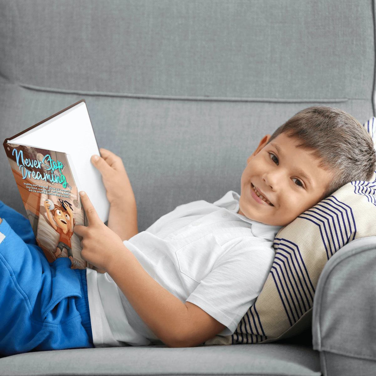 A smiling boy reads Never Stop Dreaming on a gray sofa.