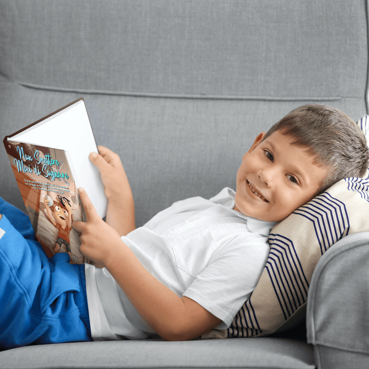 Un bambino sorridente legge Non Smettere mai di Sognare su un divano grigio.